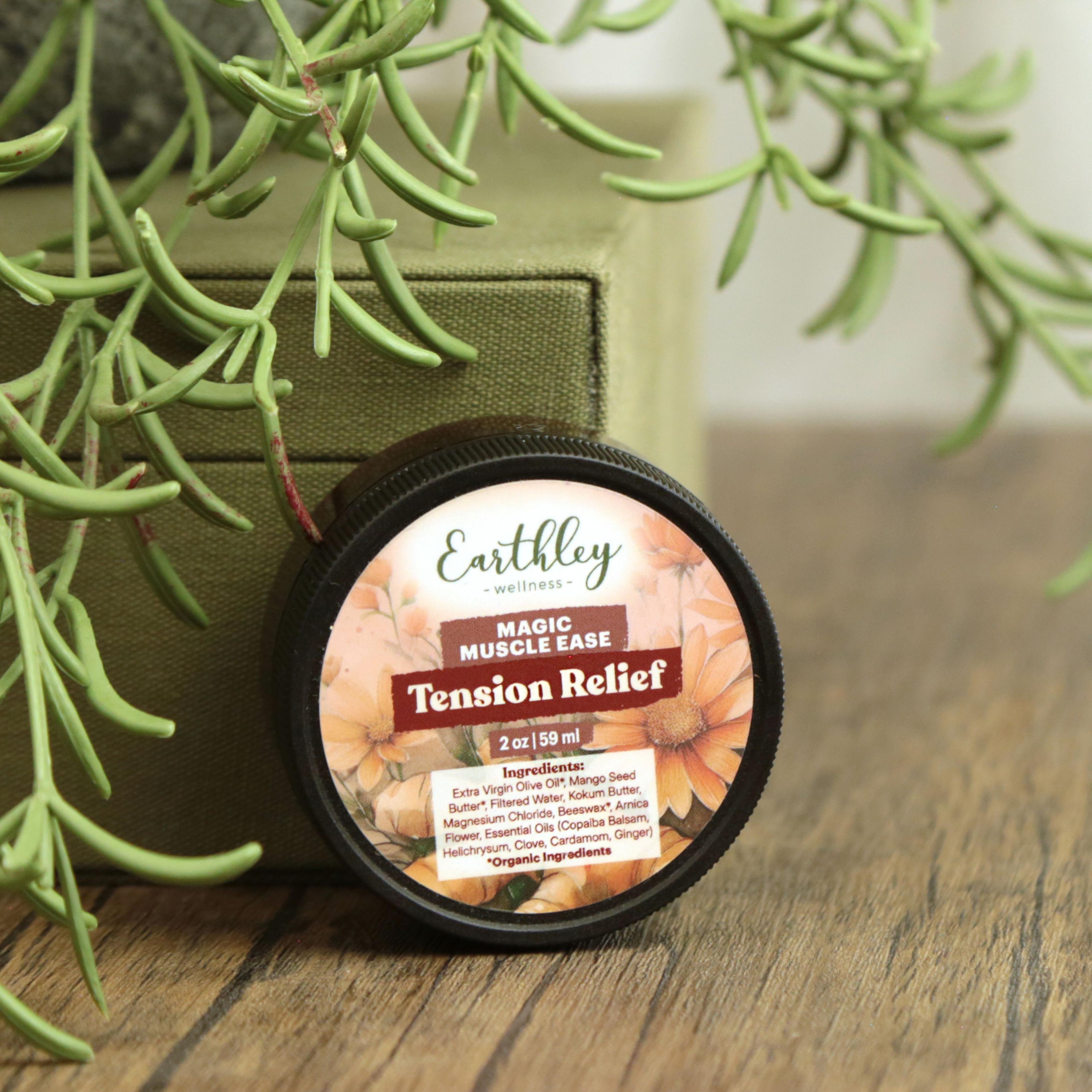 Earthley Magic Muscle Ease Tension Relief 2 oz round tin with floral label featuring orange daisy flowers, showing ingredients list on lid, on wooden surface with green box and succulent plant in background