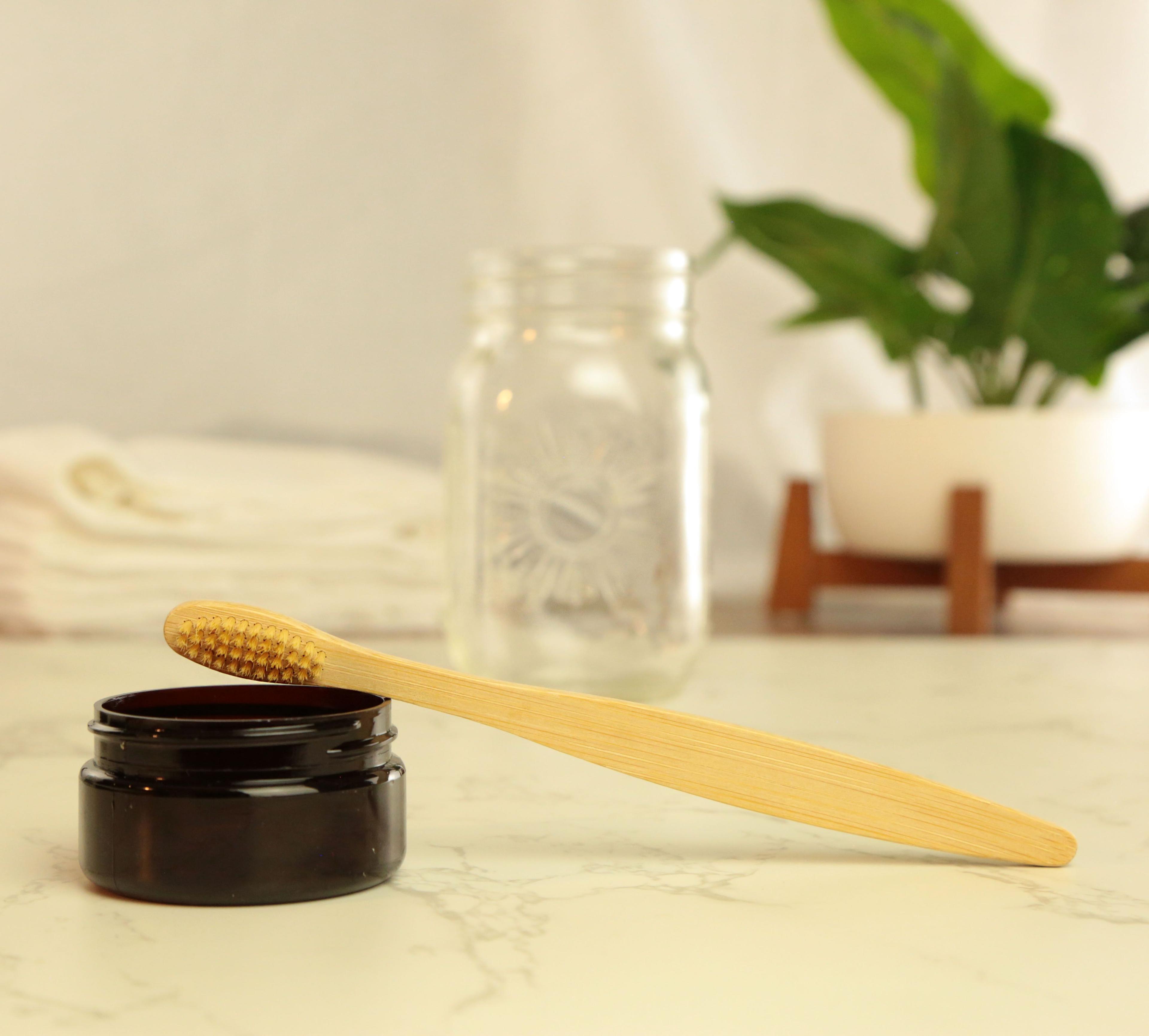 Earthley Moso Bamboo Toothbrush with natural bristles resting on open amber jar on marble surface with glass mason jar and green potted plant in background