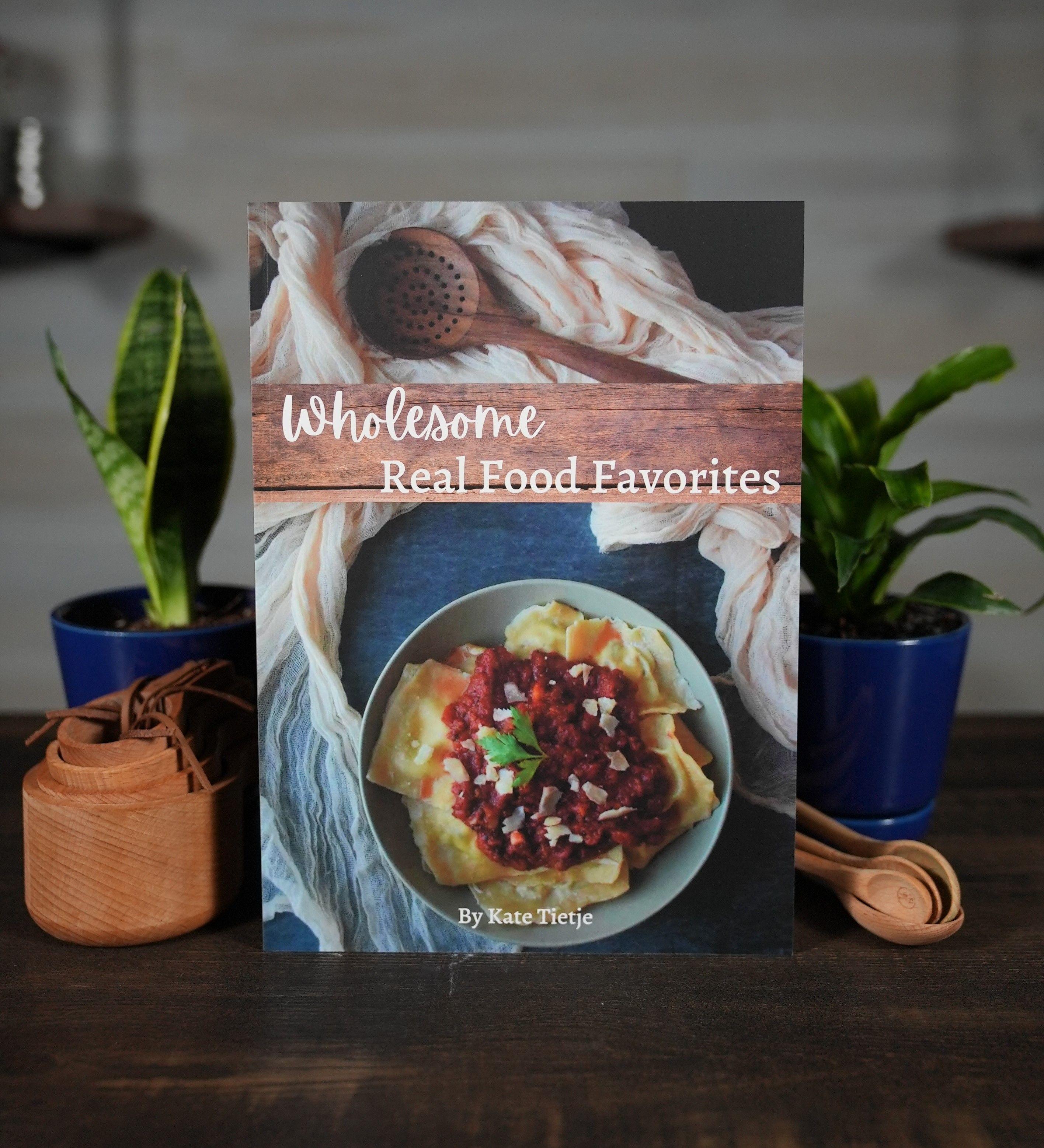 Wholesome Real Food Favorites Cookbook on table next to plants and measuring cups