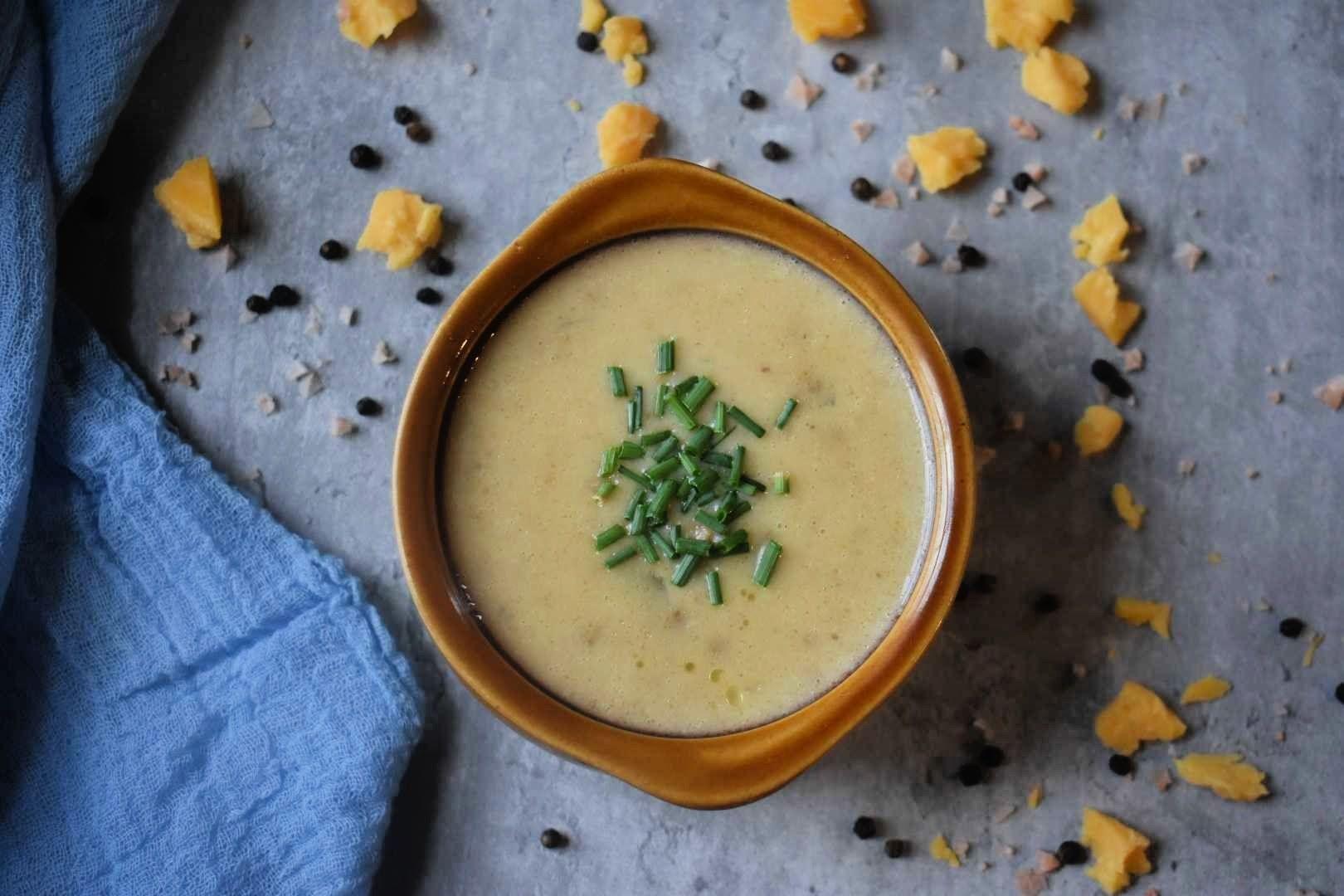 Creamy soup in brown bowl garnished with fresh chives, surrounded by scattered herbs and spices on marble surface