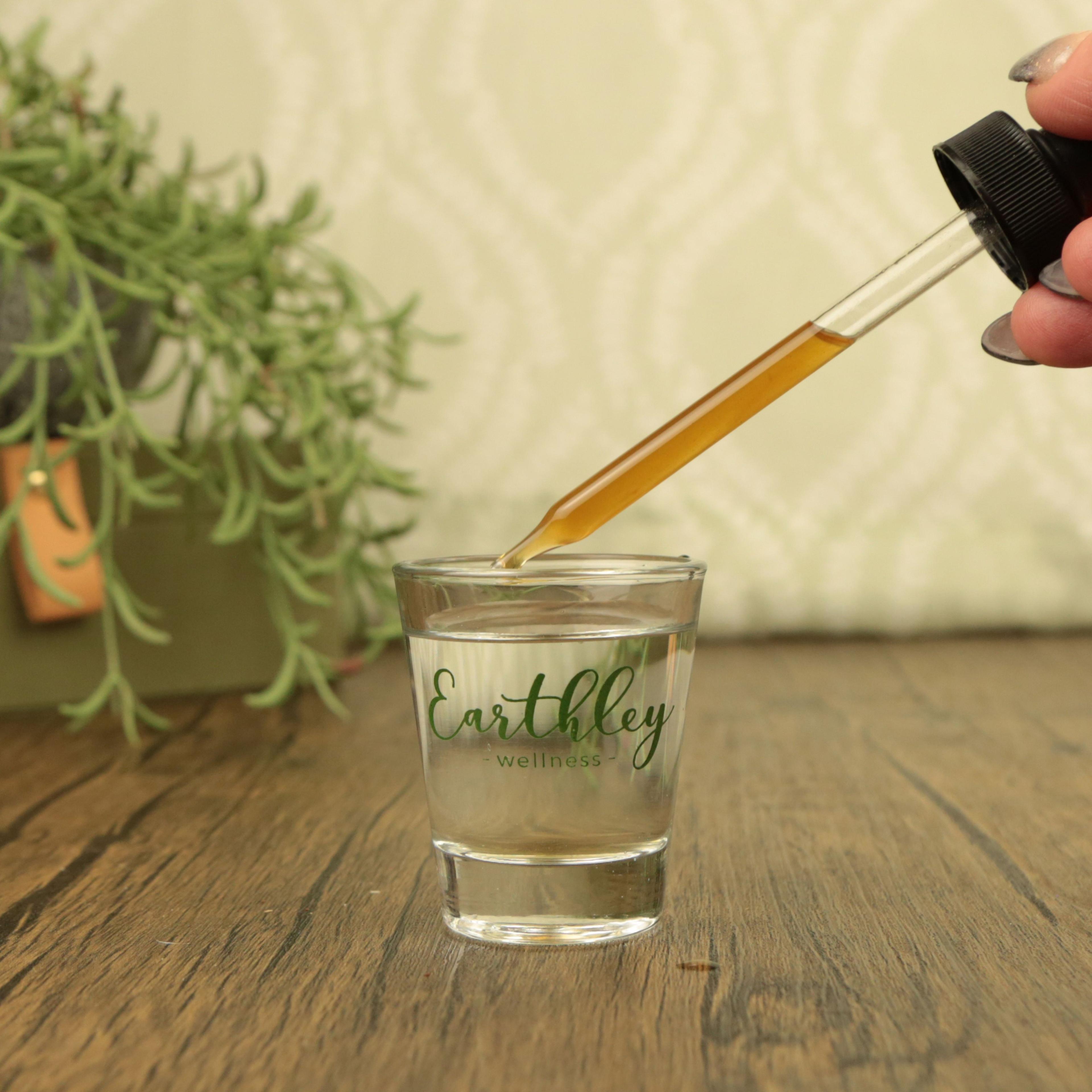 Hand dispensing golden herbal tincture from dropper bottle into Earthley Wellness branded glass on wooden surface with plant
