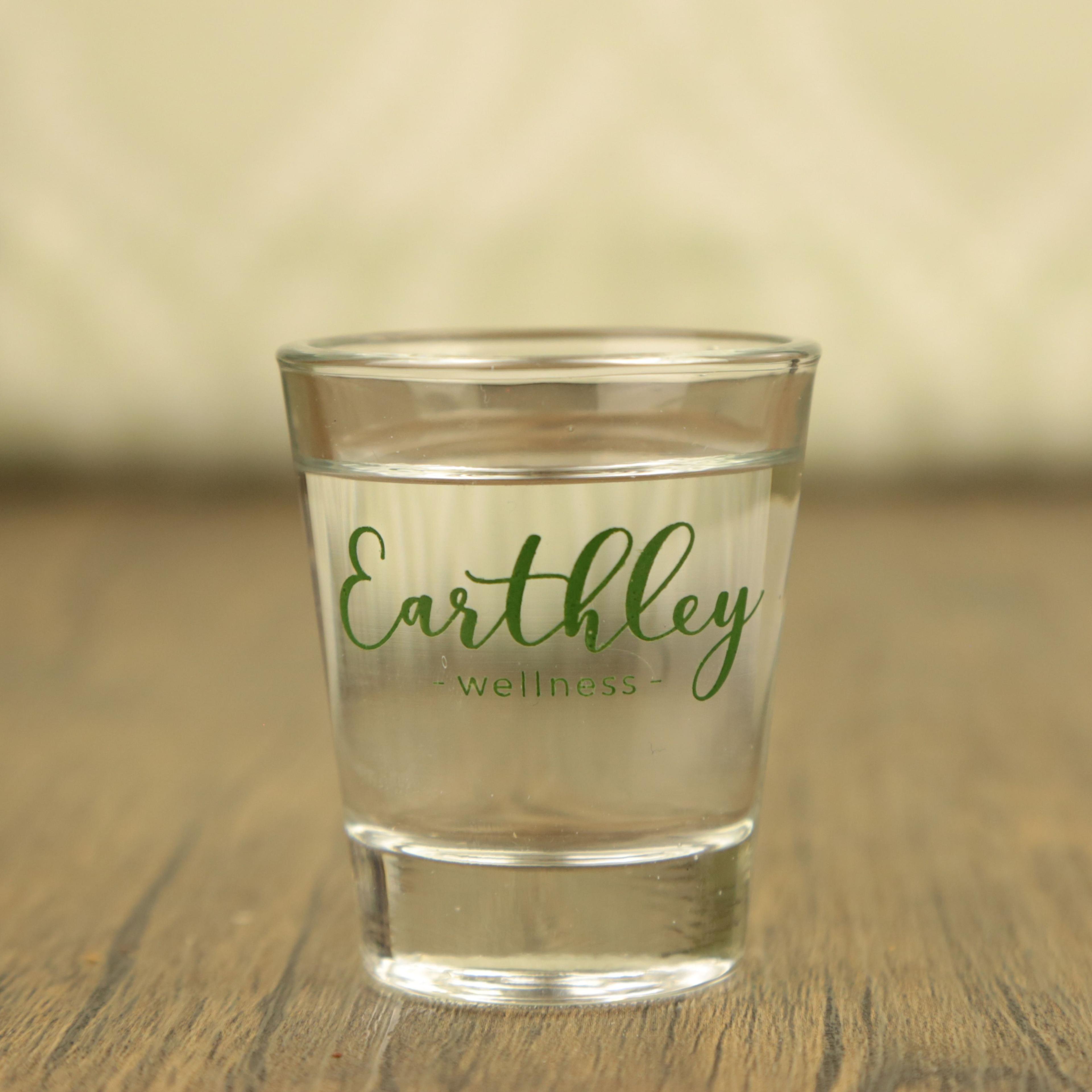 Clear glass tumbler with green Earthley Wellness logo on wooden surface