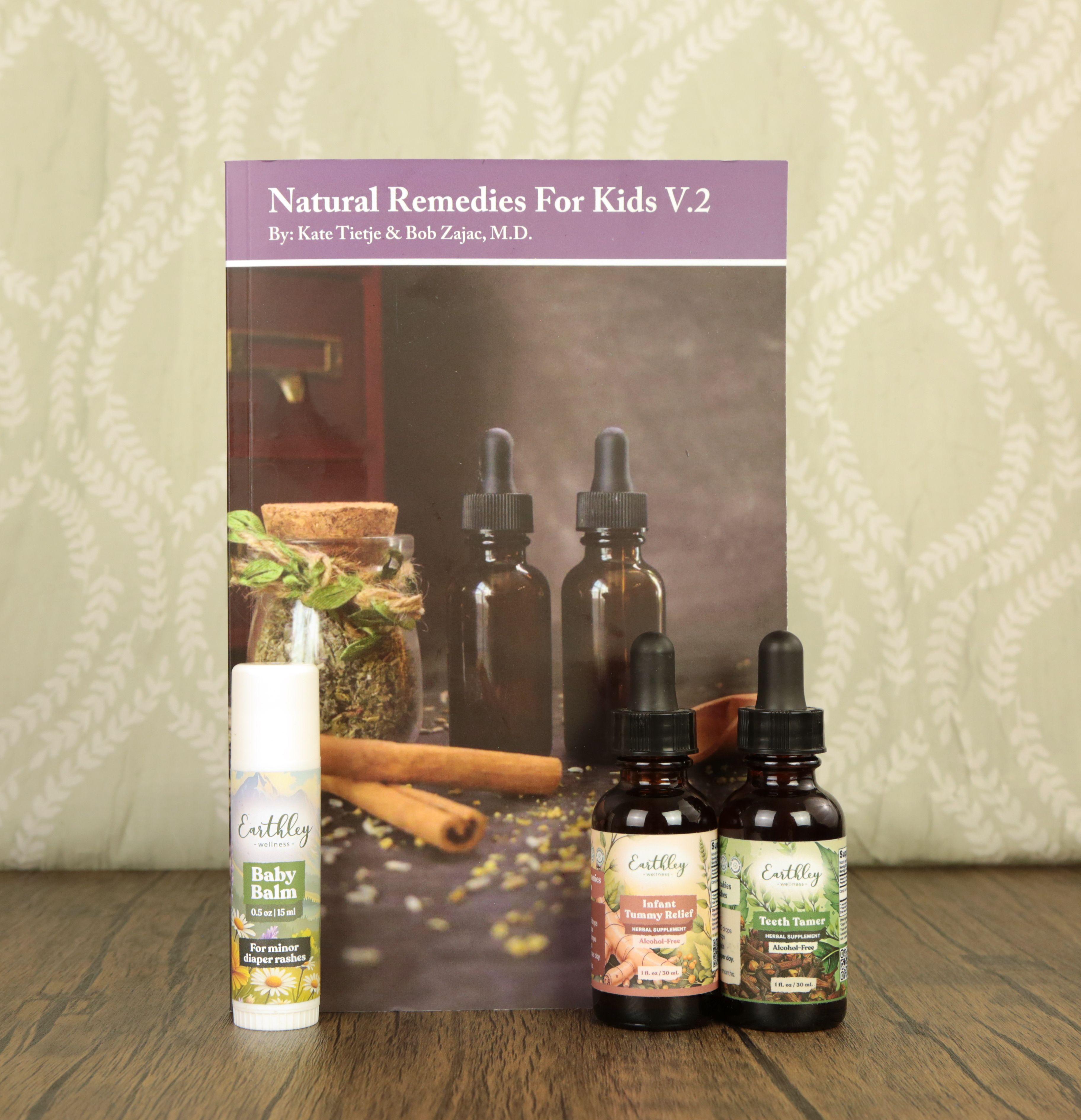 Earthley New Baby Bundle showing Natural Remedies For Kids V.2 book by Kate Tietje and Bob Zajac M.D. with Baby Balm push-up tube, Infant Tummy Relief, and Teeth Tamer amber dropper bottles on rustic wood surface with sage green fabric backdrop