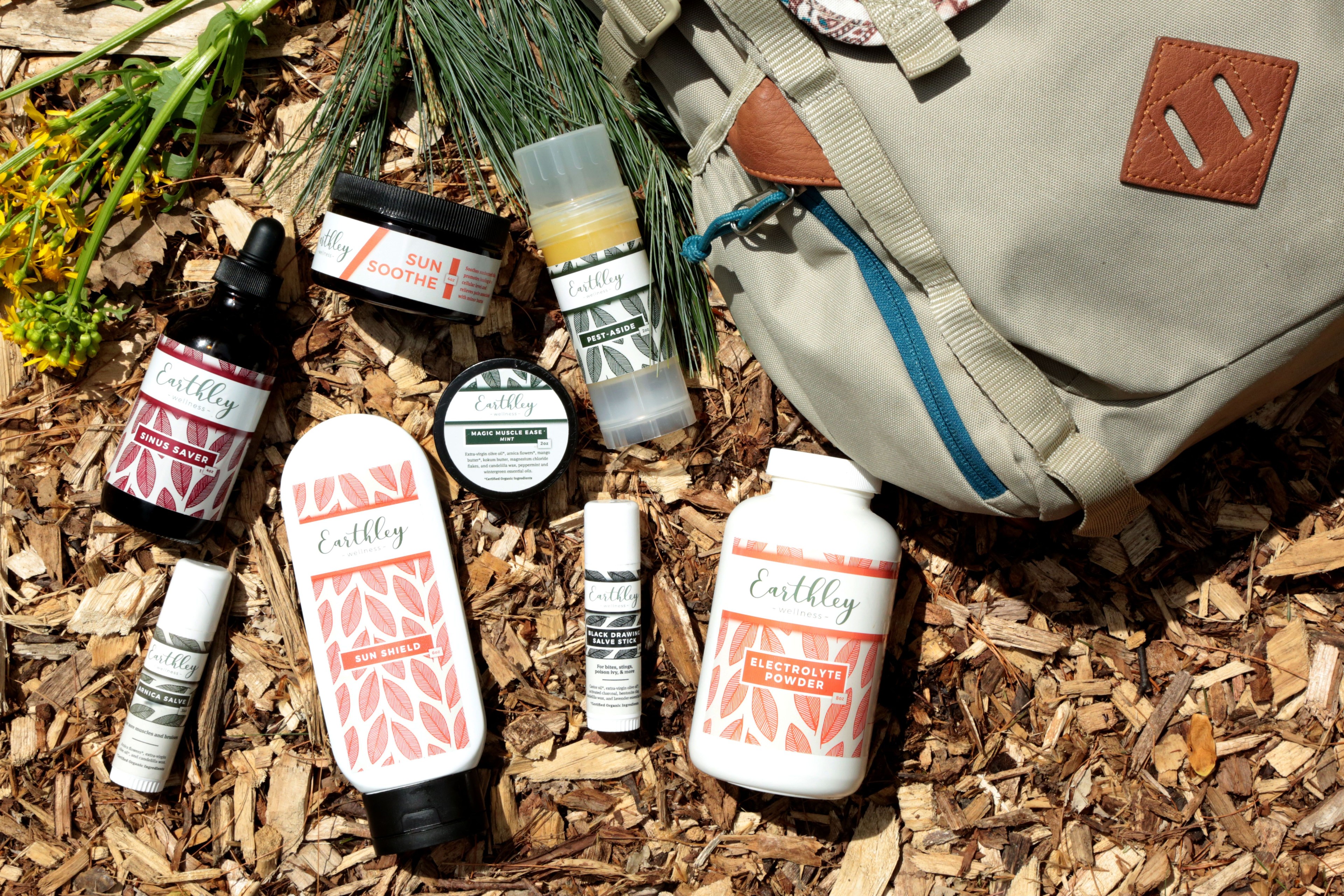 Summer collection product flatlay showing amber glass tincture bottle white jars with burgundy red leaf labels yellow lip balm tubes tan canvas backpack with brown leather patch on brown wood chip ground with green pine needles
