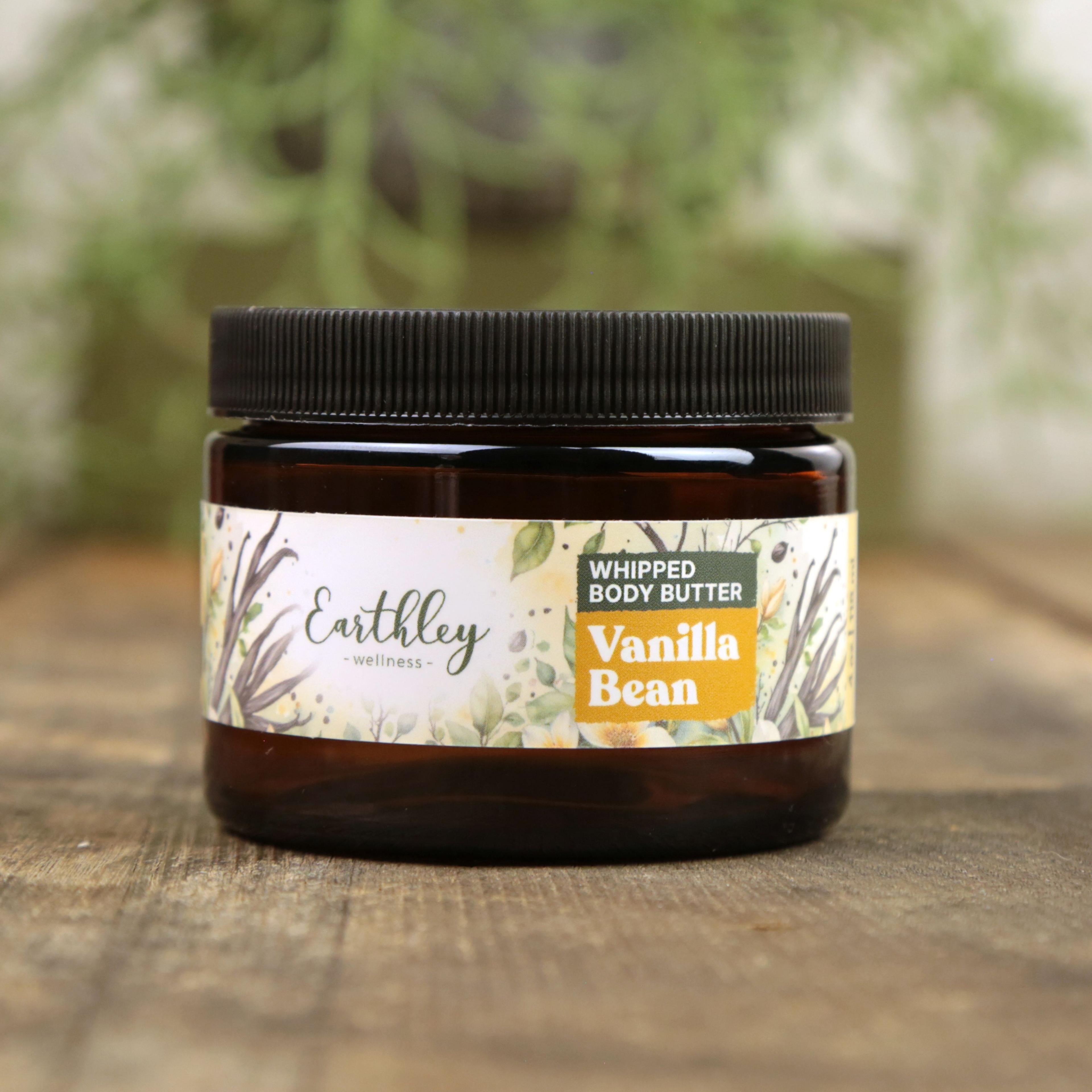 Earthley Whipped Body Butter Vanilla Bean in amber glass jar with black lid and botanical label featuring vanilla beans and eucalyptus leaves on wooden surface with greenery background
