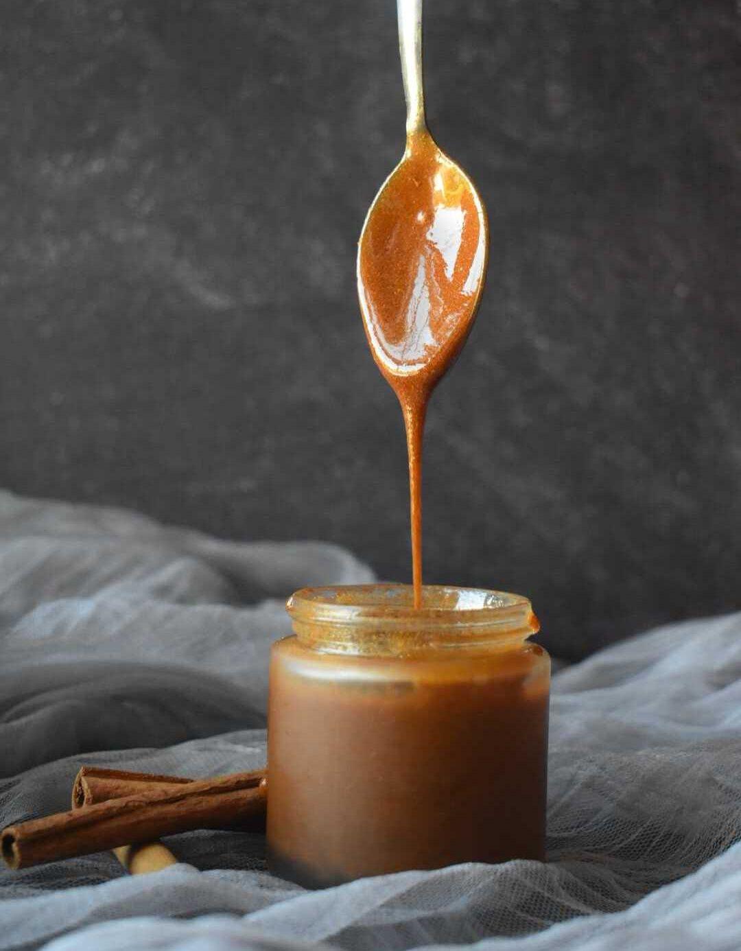 Golden honey dripping from wooden spoon into glass jar, with cinnamon sticks on gray linen fabric