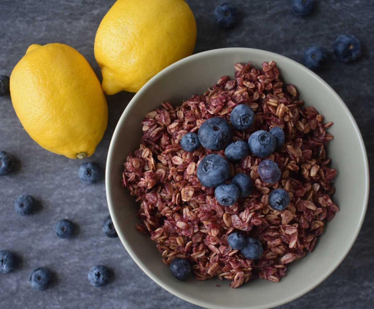 White bowl filled with purple granola topped with fresh blueberries, surrounded by lemons and scattered blueberries on dark surface