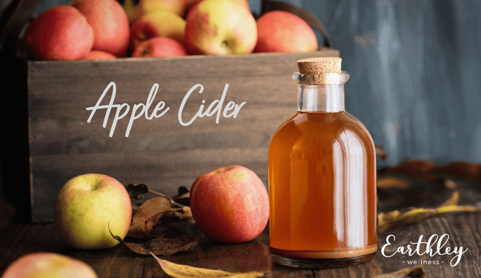 Apple Cider recipe header showing glass bottle filled with golden amber apple cider surrounded by red and green apples in wooden crate with autumn leaves on dark rustic background