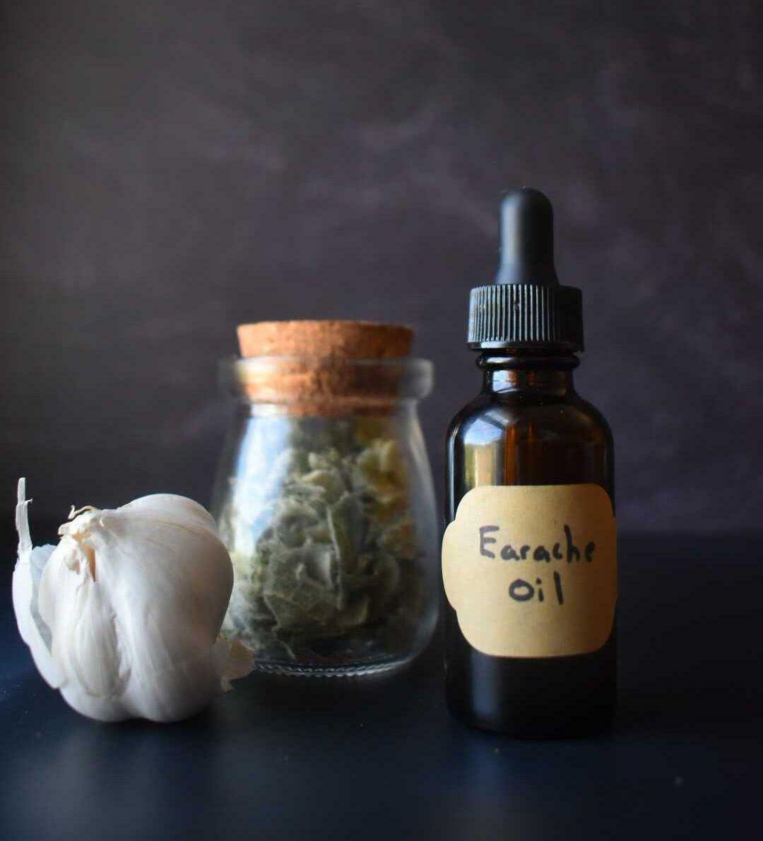 Amber dropper bottle labeled "Earache Oil" next to glass jar with dried herbs and fresh garlic bulb on dark surface