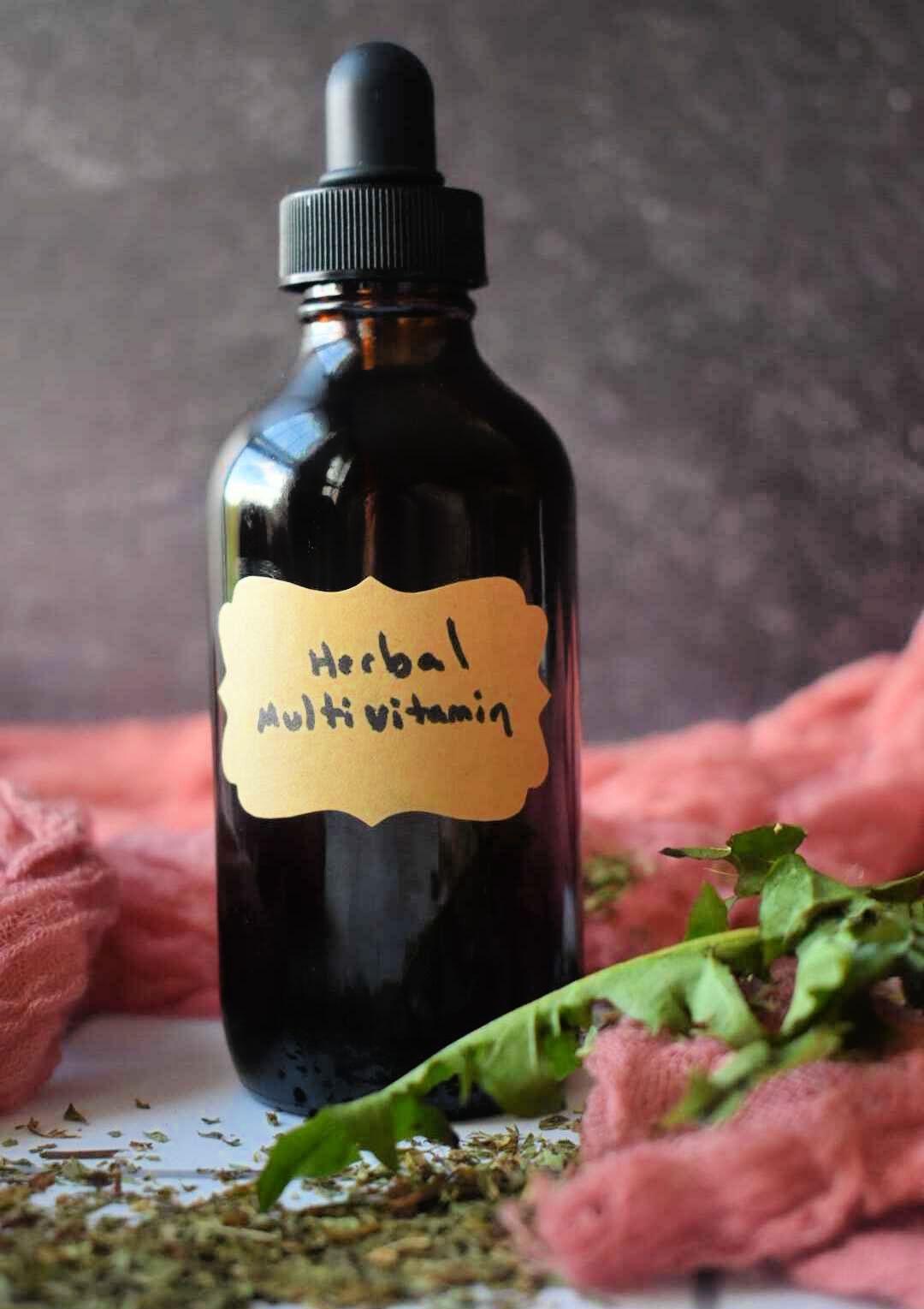 Dark amber glass dropper bottle labeled "Herbal Multi Vitamin" surrounded by fresh green herbs and coral fabric