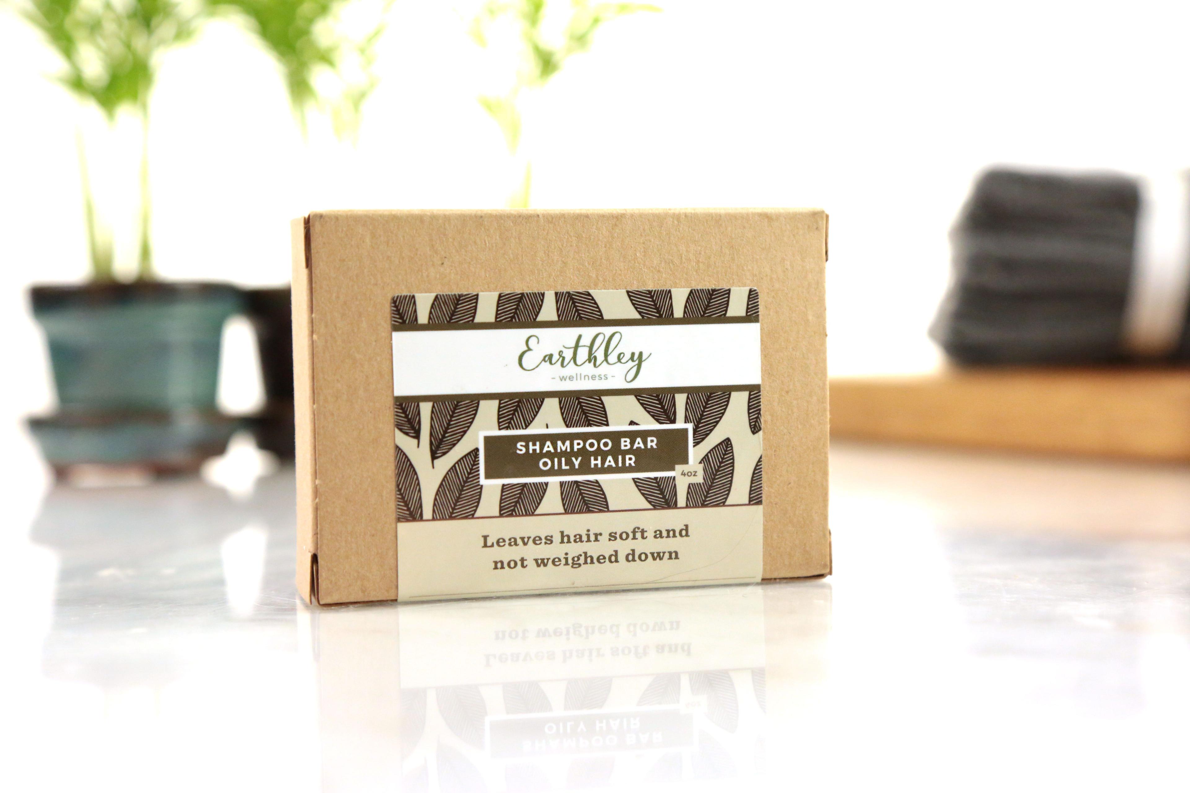 Earthley Shampoo Bar Oily Hair 4oz tan kraft box with brown leaf pattern and dark olive label on reflective white marble surface with green plant in teal pot and gray towels in background