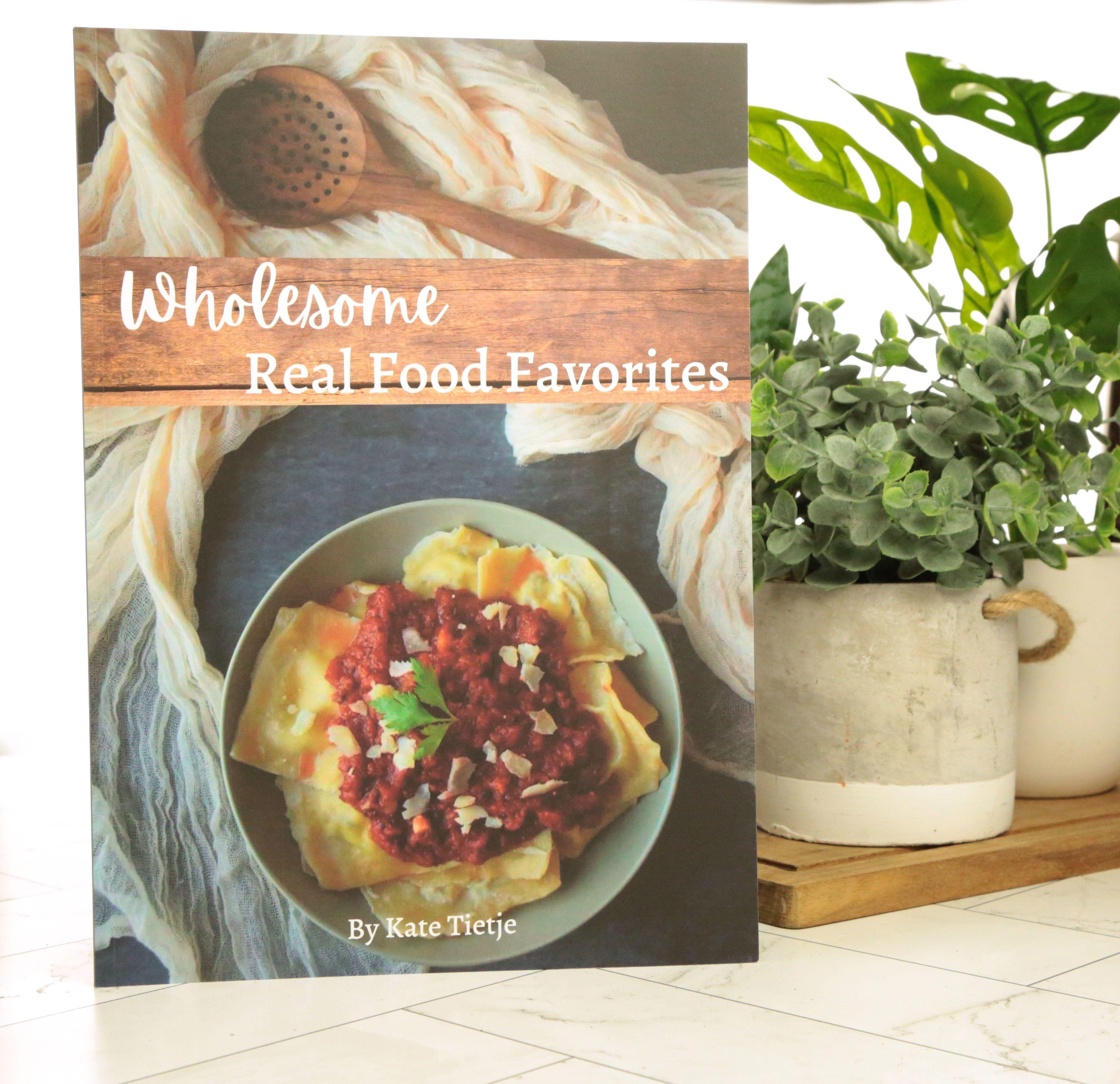 Wholesome Real Food Favorites cookbook cover by Kate Tietje featuring yellow homemade ravioli topped with dark red tomato sauce and white cheese shavings in gray ceramic bowl on blue gray surface with cream gauze fabric and dark wooden spoon with green monstera plant in cream ceramic pot beside