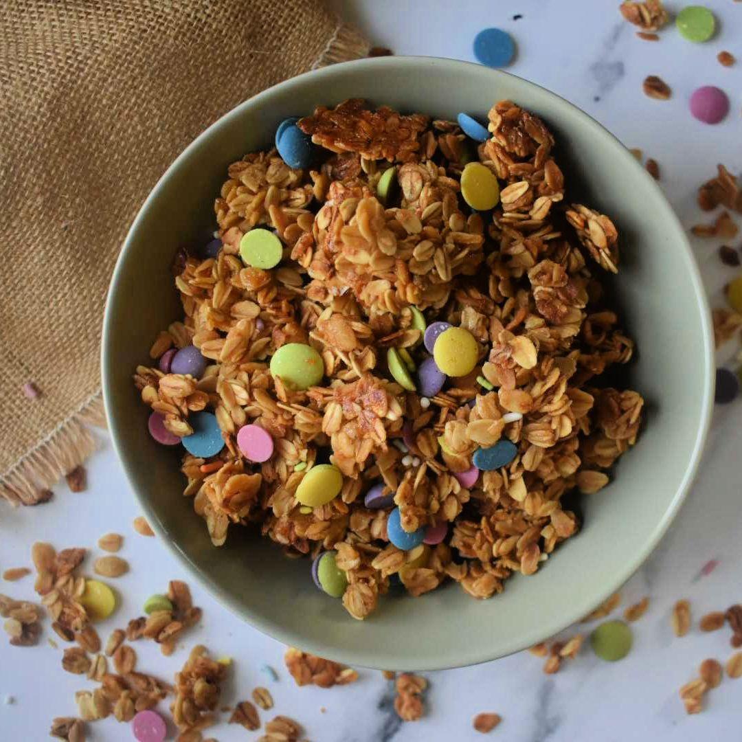 Homemade granola with oats, nuts, and colorful candy pieces in white bowl on marble surface