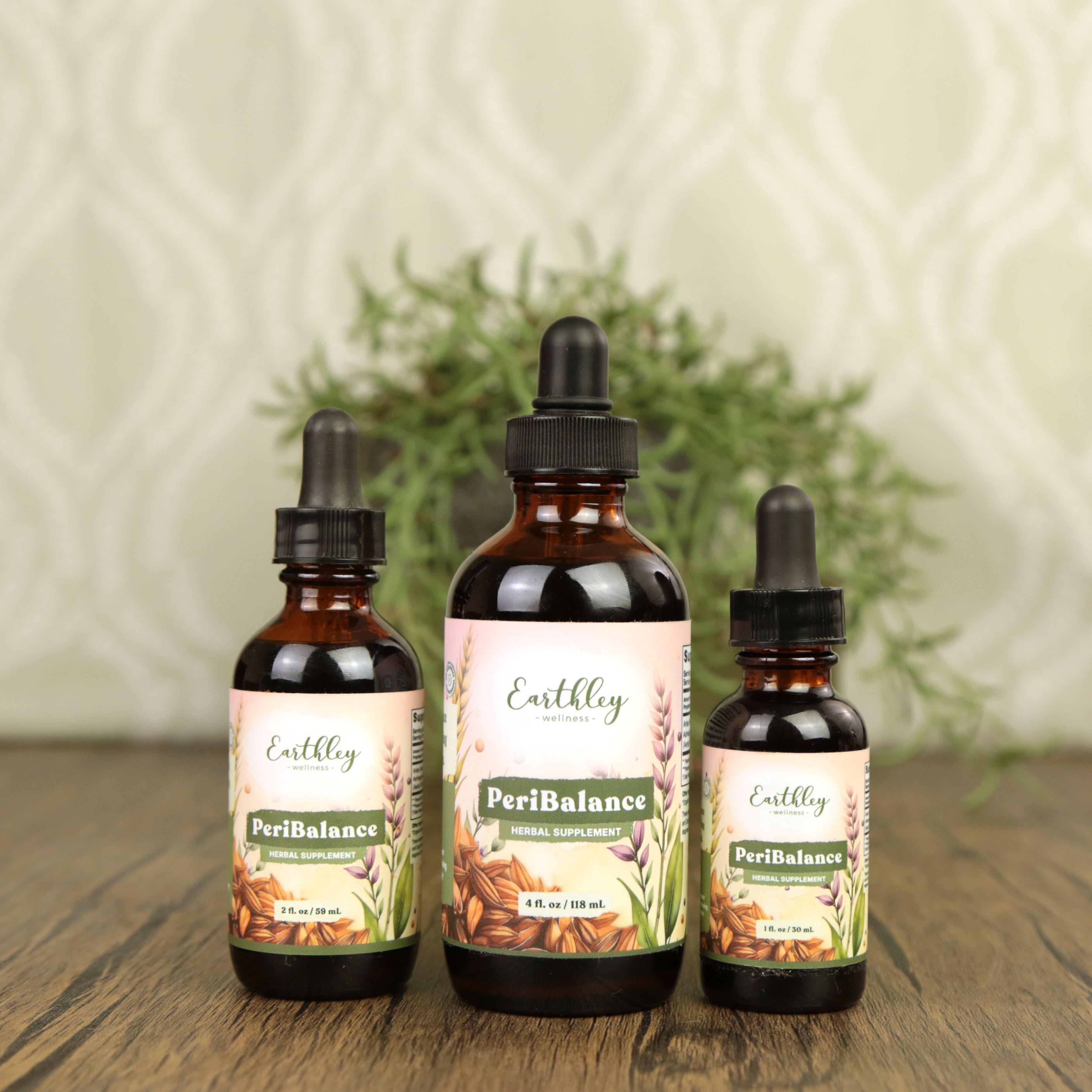 Three Earthley PeriBalance amber dropper bottles in 1 oz, 2 oz, and 4 oz sizes with soft pink botanical labels featuring oats and wheat arranged on wooden surface with greenery in background