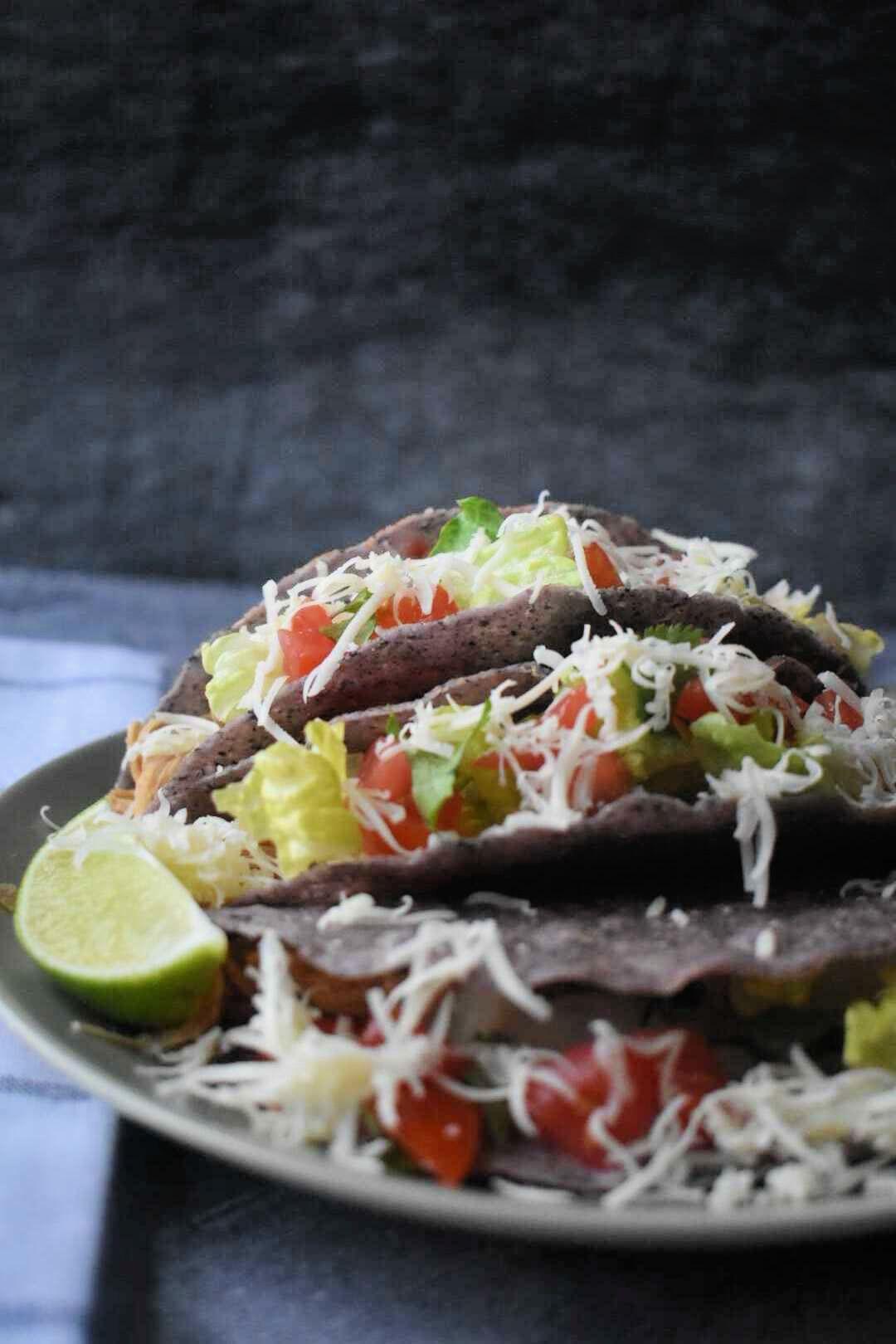 Blue corn tortilla tacos filled with lettuce, tomatoes, cheese and lime wedges on white plate